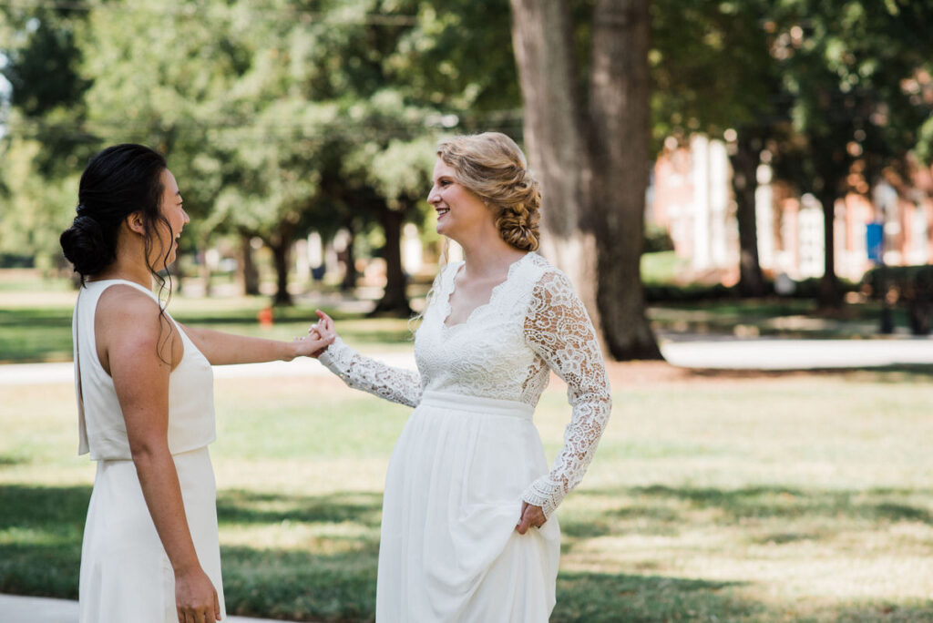 Two women, one in a sleeveless white gown and the other in a long-sleeved lace wedding dress, smiling and holding hands in a sunny park