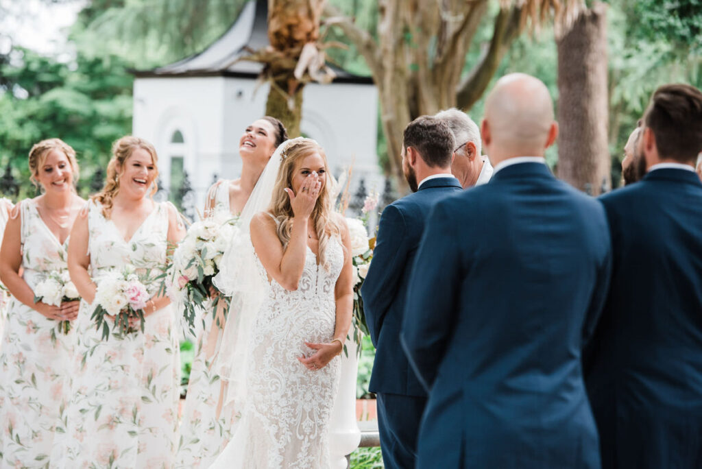 A joyful wedding scene with the bride laughing and covering her mouth, surrounded by bridesmaids in patterned dresses, and groomsmen in the background facing them