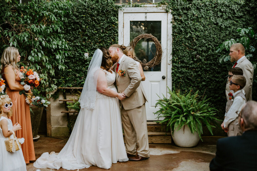 A bride and groom sharing a kiss on the lips, with guests in semi-formal attire looking on, in front of a door framed by lush greenery and a wreath.