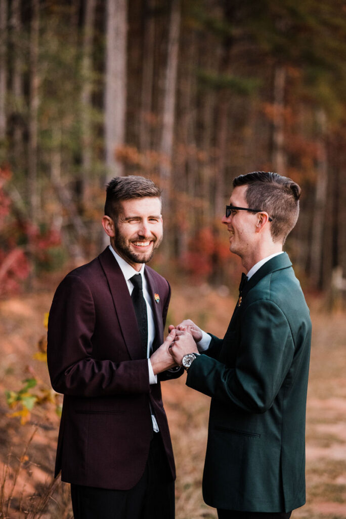 Two grooms smiling and holding hands in a forest, one dressed in a burgundy suit and the other in a green suit, with autumn leaves in the background