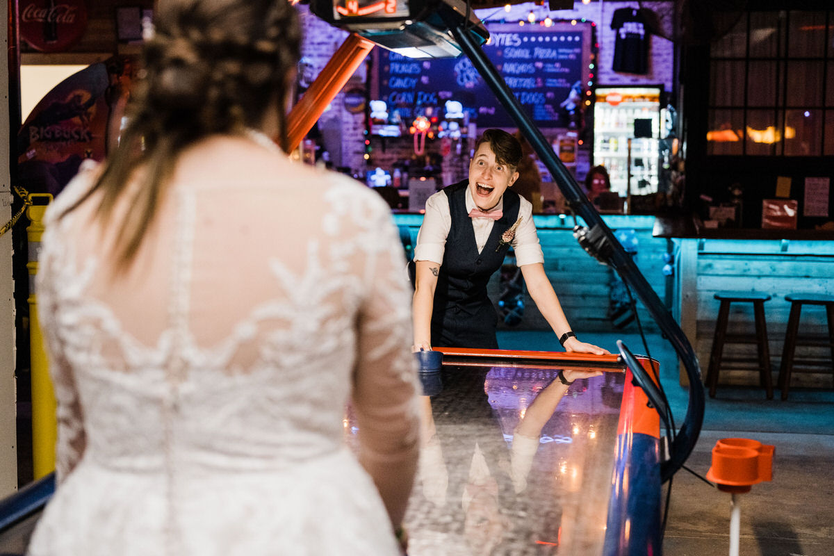 A non-traditional wedding theme in an arcade with a bride in a detailed wedding gown playing air hockey with a surprised-looking woman in a vest and bow tie at a colorful, neon-lit arcade