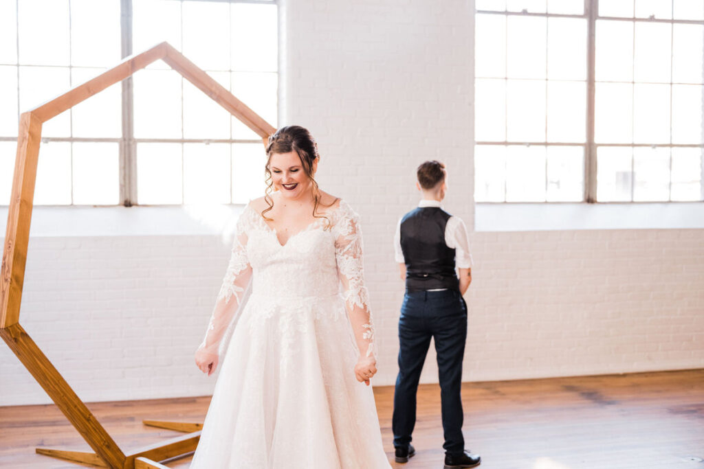 Bride in a lace wedding gown smiling widely, with her back towards a person in suspenders, in a bright room with large windows.