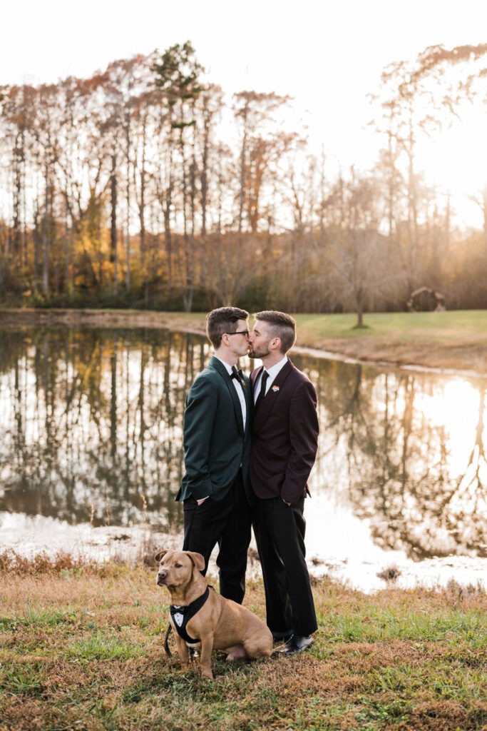 A couple kissing by a serene lake with a reflection of trees in the water, accompanied by a dog wearing a bow tie