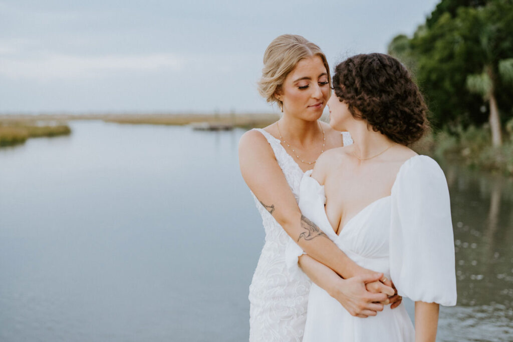 Two brides embracing near a tranquil water body, one with a tattooed arm