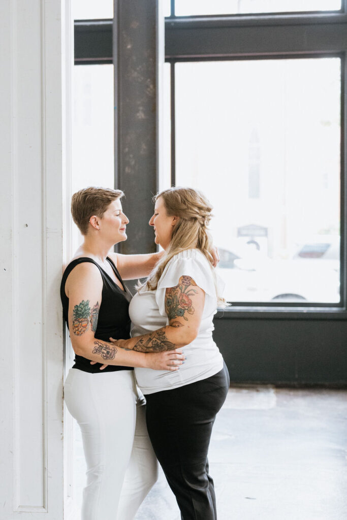 Two women embracing and smiling at each other by a bright window, showing off colorful arm tattoos, in a warmly lit industrial-style room.