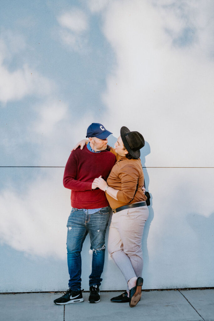 A couple playfully touching foreheads with a clear blue sky in the background, man in a red sweater and baseball cap, woman in a hat and light brown jacket, creating a relaxed and affectionate atmosphere.