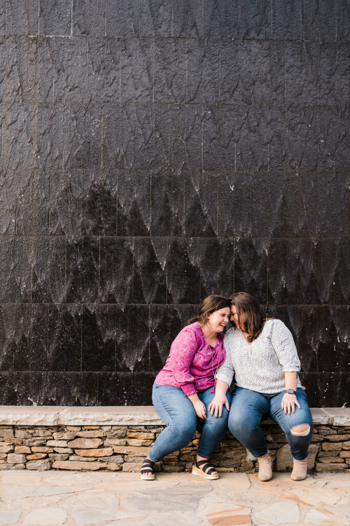 Two women laughing and sitting closely on a stone ledge, with a dark textured wall behind them, exuding joy and closeness in casual attire.