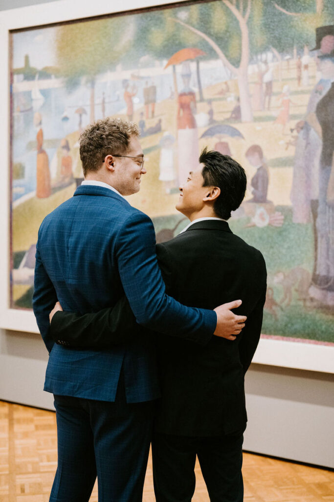 Two men in formal wear, one in a blue suit and the other in black, smile at each other, with a large Seurat-like pointillist painting in the background.