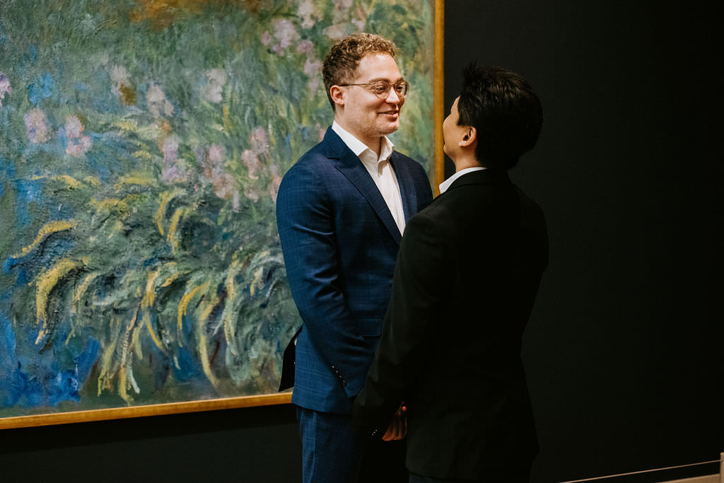 Against the backdrop of a large, textured Monet-style water lily painting, the man in the blue suit, with glasses, shares a joyful moment with his partner.