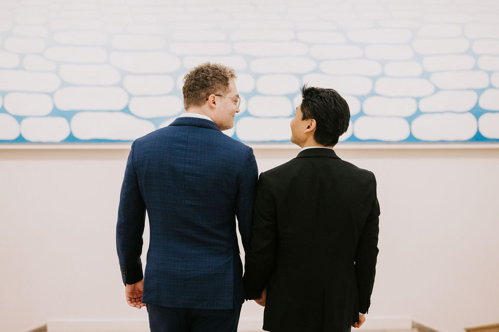 View from behind two men in suits standing contemplatively before an abstract painting with a pattern of blue cloud-like shapes on a white background.