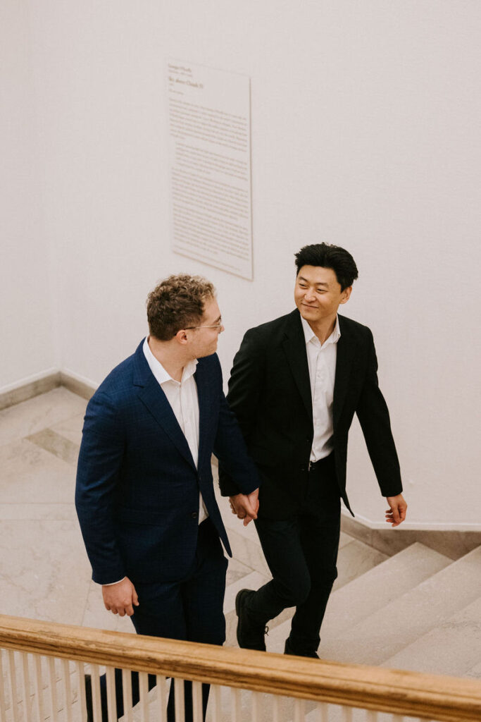 Walking up a classic museum staircase, the men are engaged in conversation, with one looking back and smiling, emphasizing their shared connection.