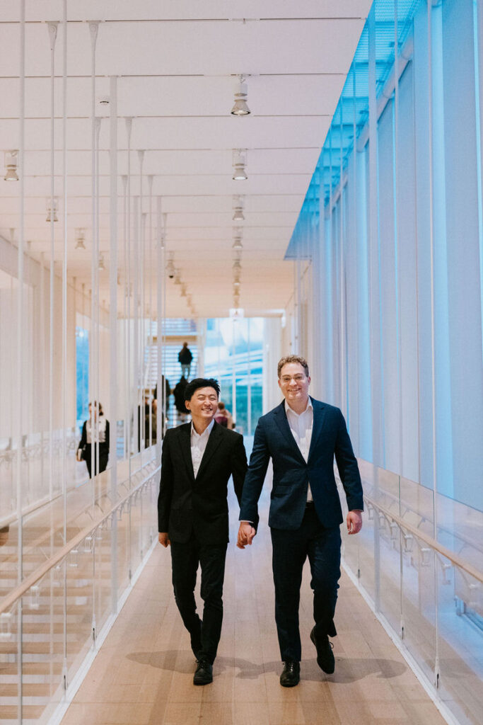 A cheerful stroll through a bright museum corridor, both men looking ahead, hand in hand, displaying a sense of companionship and ease.