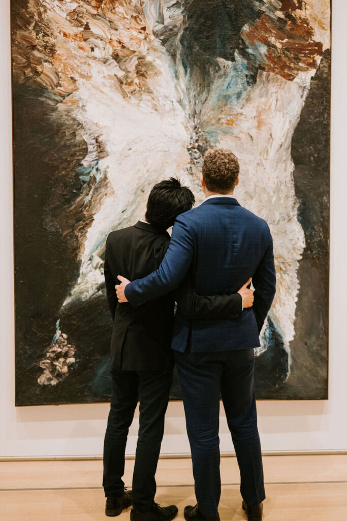 Intimate moment between the two men, with one resting his head on the other's shoulder, standing in front of an abstract painting with explosive white and earth tones.
