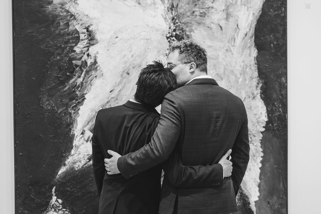 A black and white photo captures a tender moment as the two men hug, standing before an expansive abstract painting with dynamic brushstrokes.
