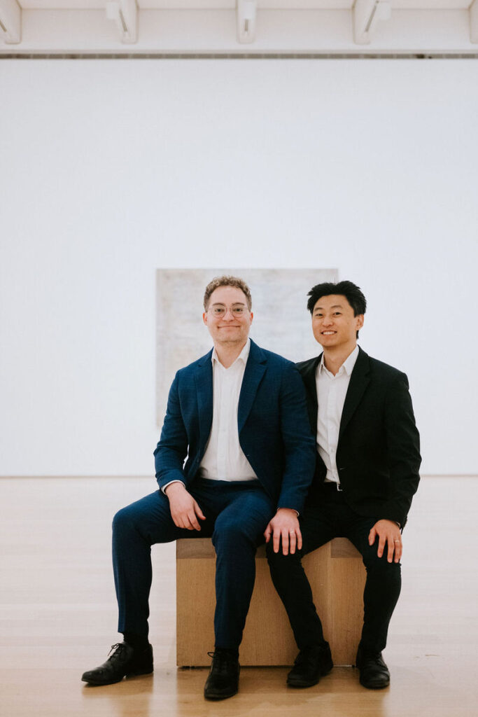 Both men, in smart casual attire, sit on a wooden block in a minimalist gallery space, with a large abstract painting in the background, sharing a moment of relaxation.