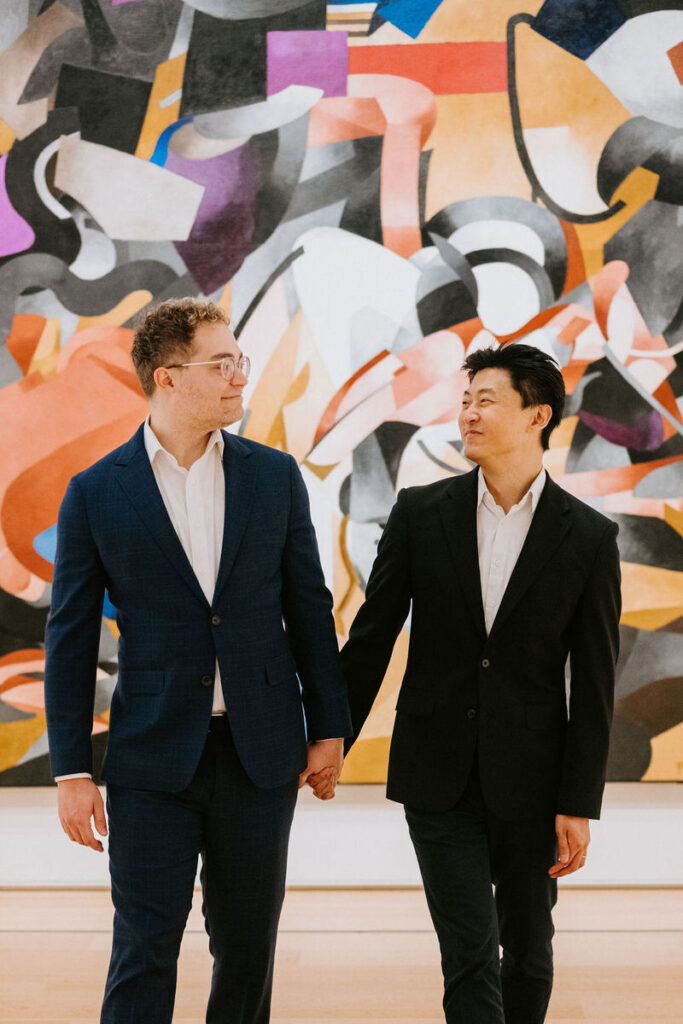 Another scene showing the two men hand-in-hand, with the man in blue looking at his partner affectionately as they stand in front of a mural with colorful geometric patterns.