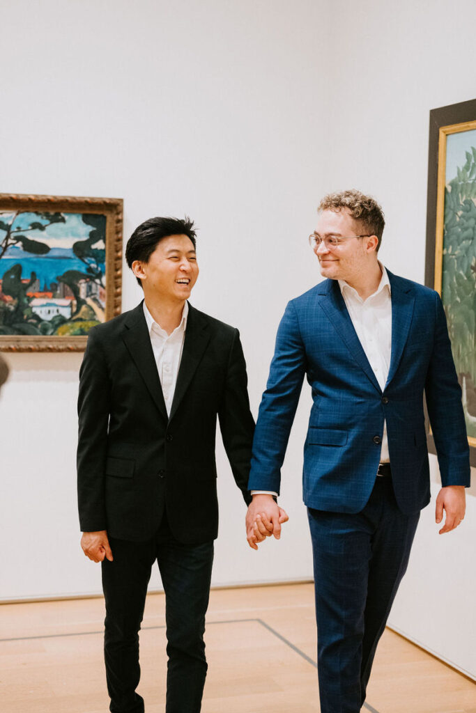 The men, now seen from the front, continue holding hands before the abstract painting, their smiles suggesting a shared joke or memory.