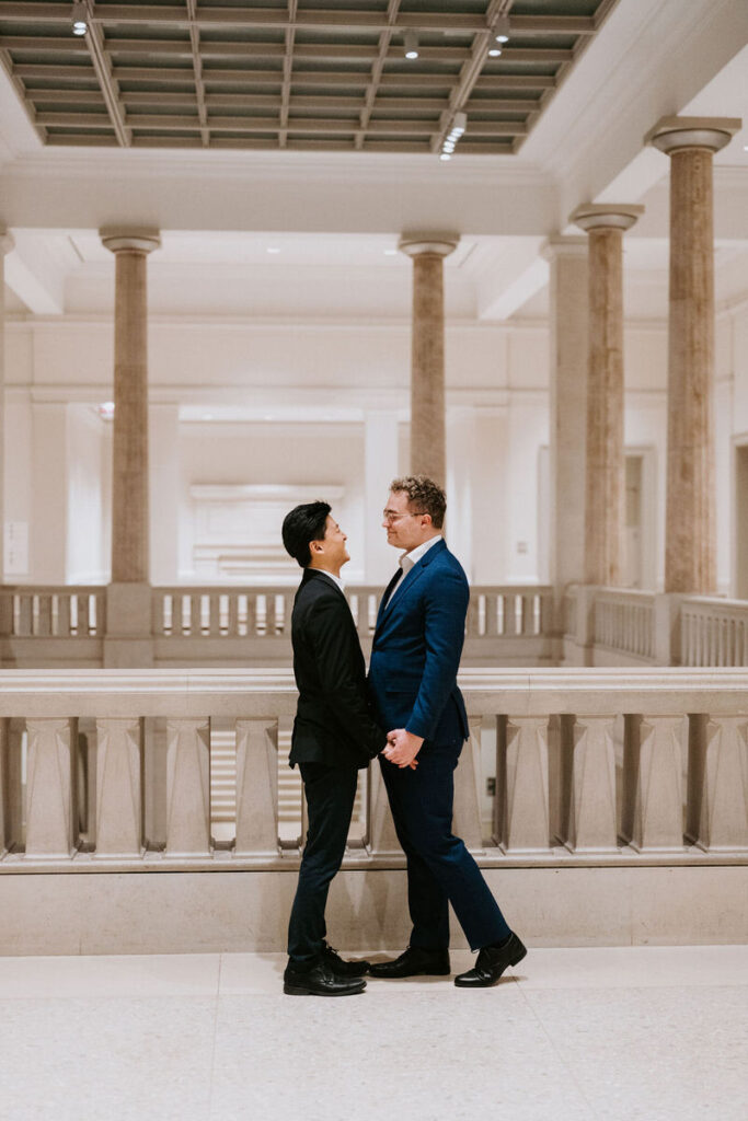 The men standing in a grand, columned hallway, facing each other, about to share a kiss, adding a personal touch to the grandeur of the architecture.