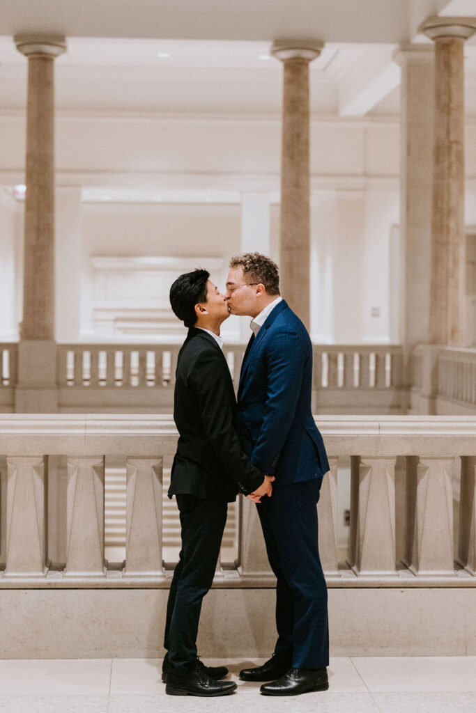 A tender moment captured as the men kiss, the classical architecture around them emphasizing the romance of their connection.