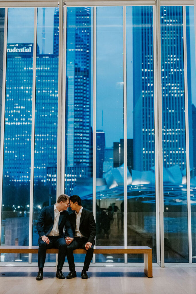 Captured in a candid moment, they sit close on a bench, their reflections mirrored in the expansive glass windows showcasing the city skyline.