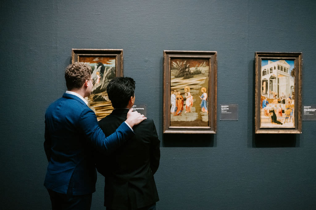 Two men in business attire viewing a painting in a dark-hued art gallery, one pointing out details on the canvas.