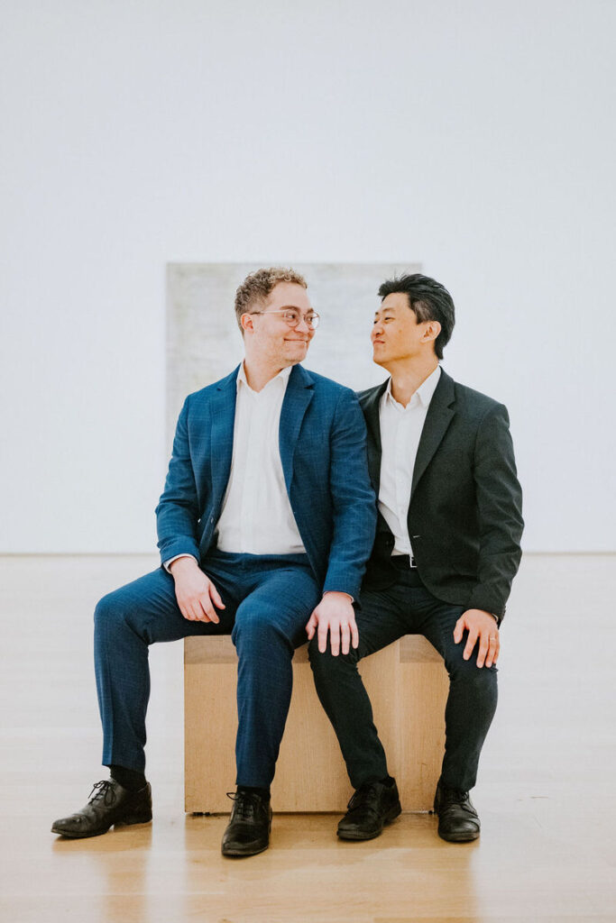 Two men sitting side by side on a bench in an art gallery, smiling and looking at each other, with a large white painting as the backdrop