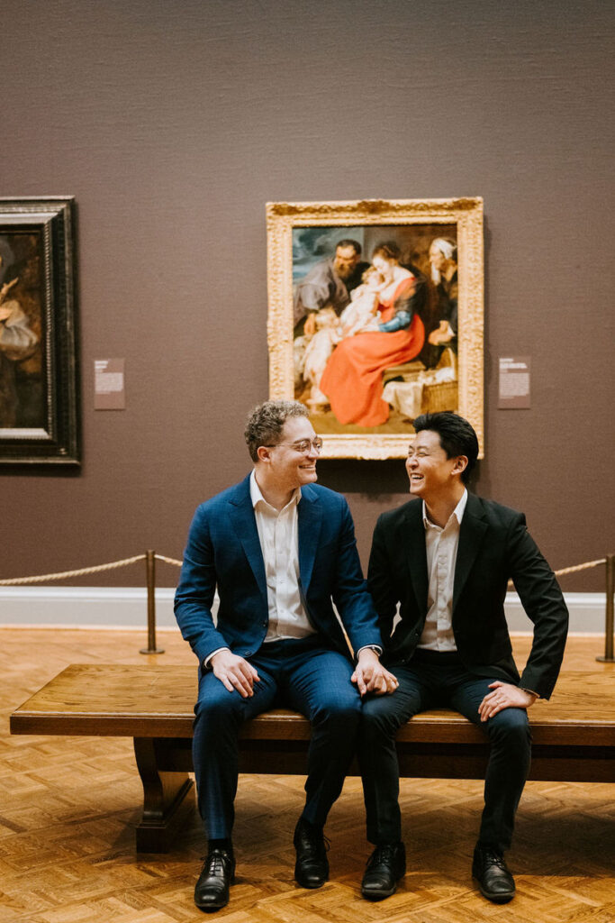 The same pair sharing a laugh while sitting on a wooden bench, with an ornate golden-framed painting in the background.