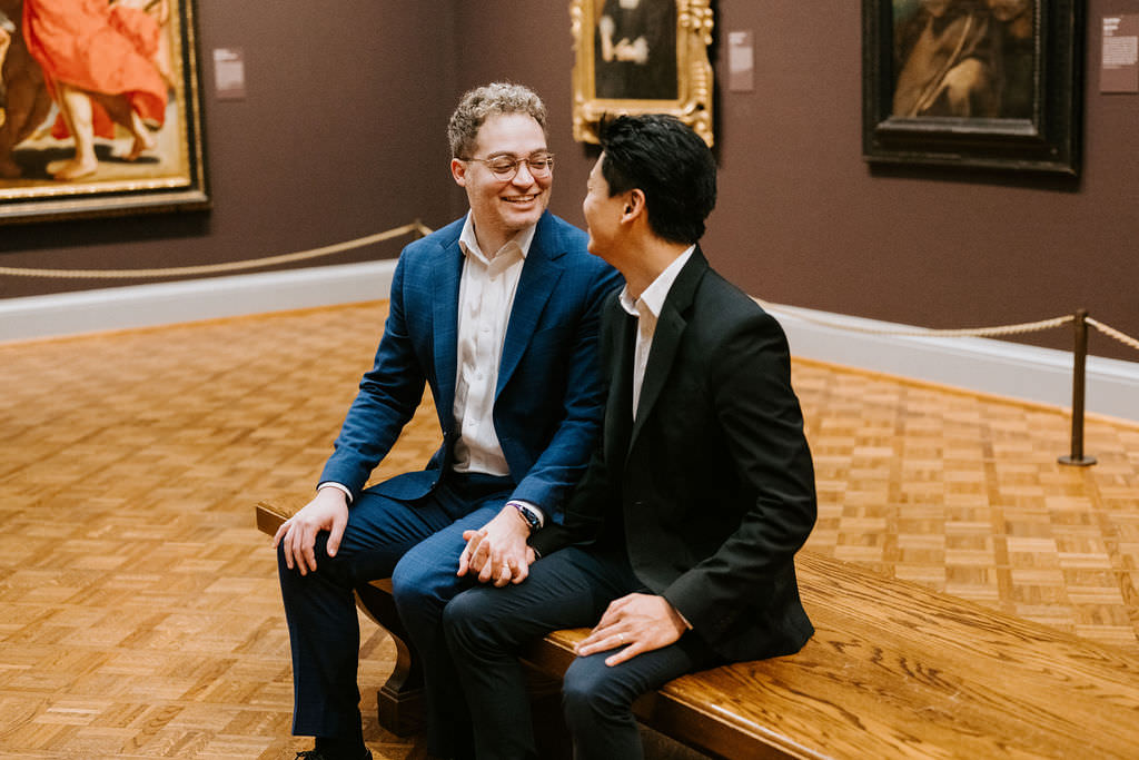 Both men smiling and conversing on a wooden bench in front of a vivid painting, enjoying each other's company.