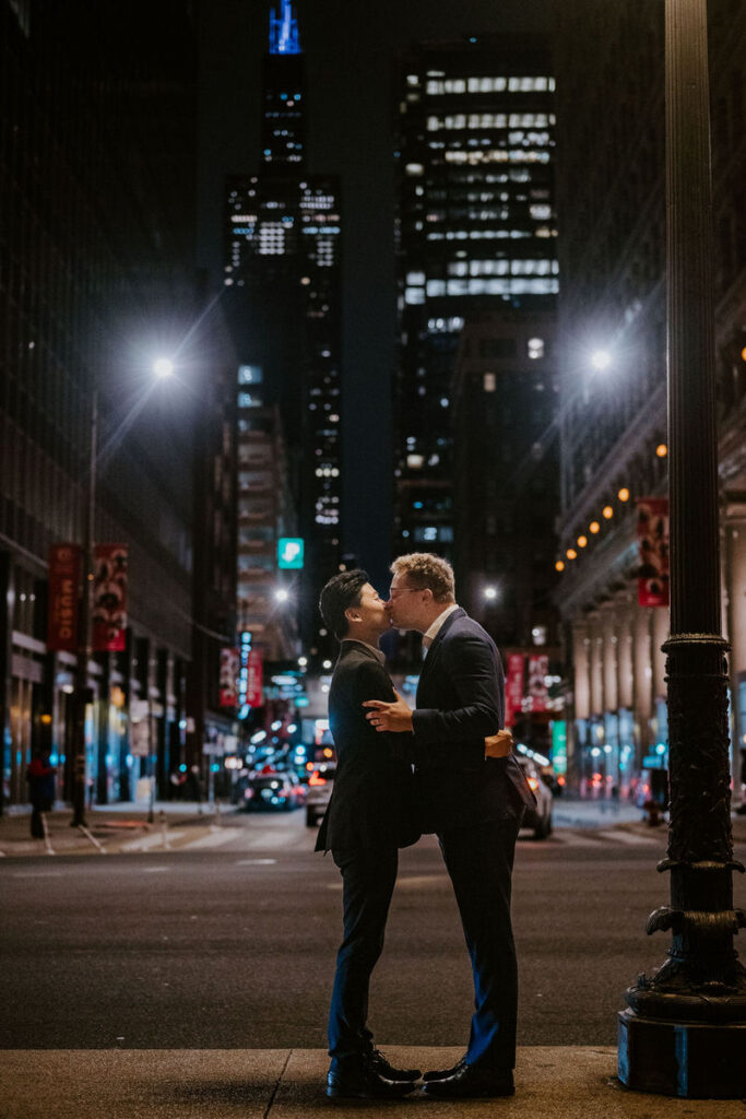 Two men standing close, sharing a tender moment under the bright city lights of Chicago at night, with a view of illuminated skyscrapers in the background.