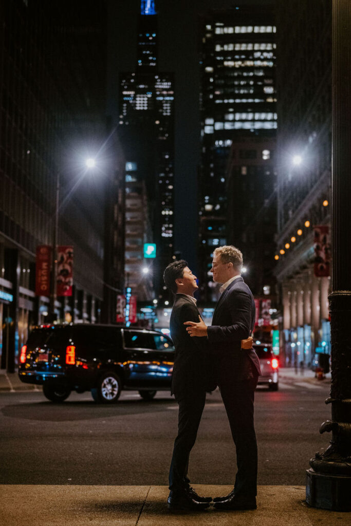 Two men holding hands, romantically engaged in a kiss on a city street at night, with the glow of Chicago's skyline and streetlights surrounding them