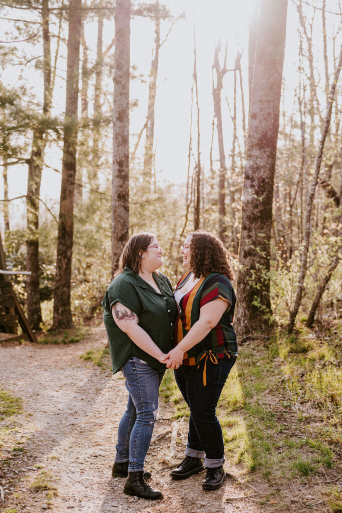 Two women holding hands and looking into each other’s eyes, one in a green shirt and the other in a striped dress, standing on a forest path, exuding warmth and affection in a peaceful setting.
