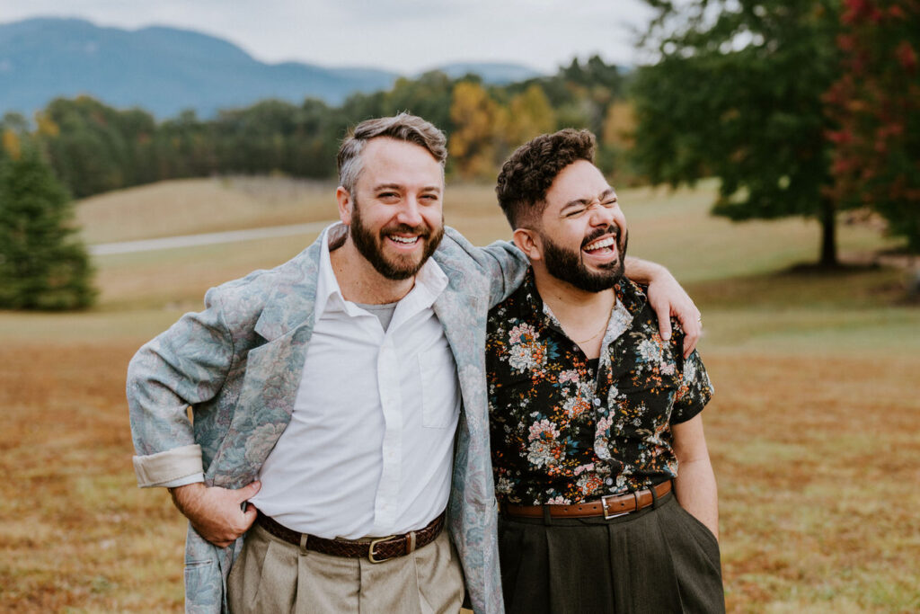 Two men laughing together in a field with trees and mountains behind them, one in a floral shirt and the other in a patterned jacket, radiating joy and camaraderie.