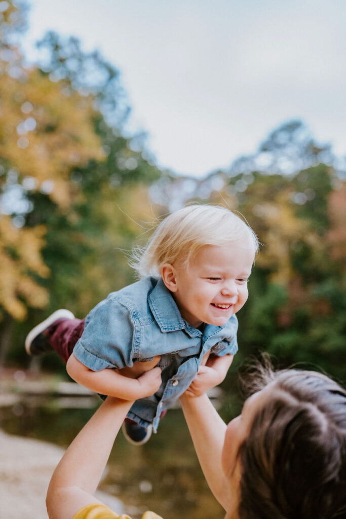 A child in a denim jacket flying in the air held by an adult, against a backdrop of autumn foliage, capturing a playful and happy moment.