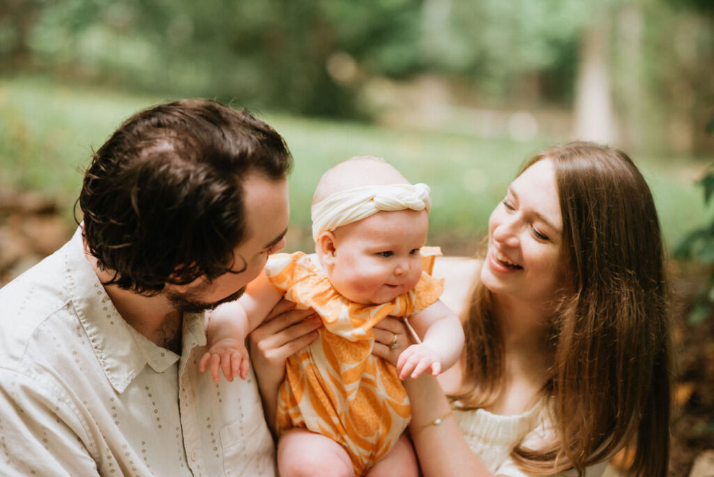 A family with a baby girl wearing an orange dress, the father kissing the baby’s cheek and the mother smiling brightly, creating a scene of parental love and happiness in nature.