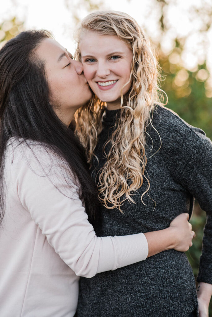 Two women closely embracing, one giving a gentle kiss on the cheek of the other, both smiling joyfully in a natural, sunlit setting, reflecting a loving and happy relationship.