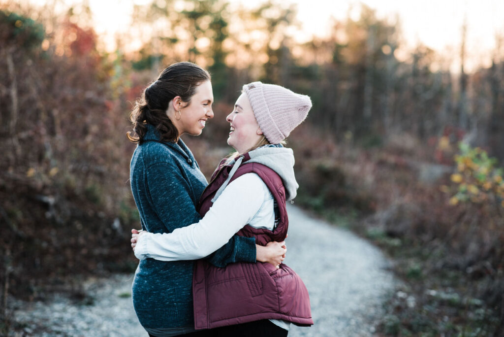 Two women wrapped in each other's arms on a chilly day, one in a pink beanie and maroon vest, the other in a blue cardigan, sharing a laugh against a backdrop of rustic fall colors.