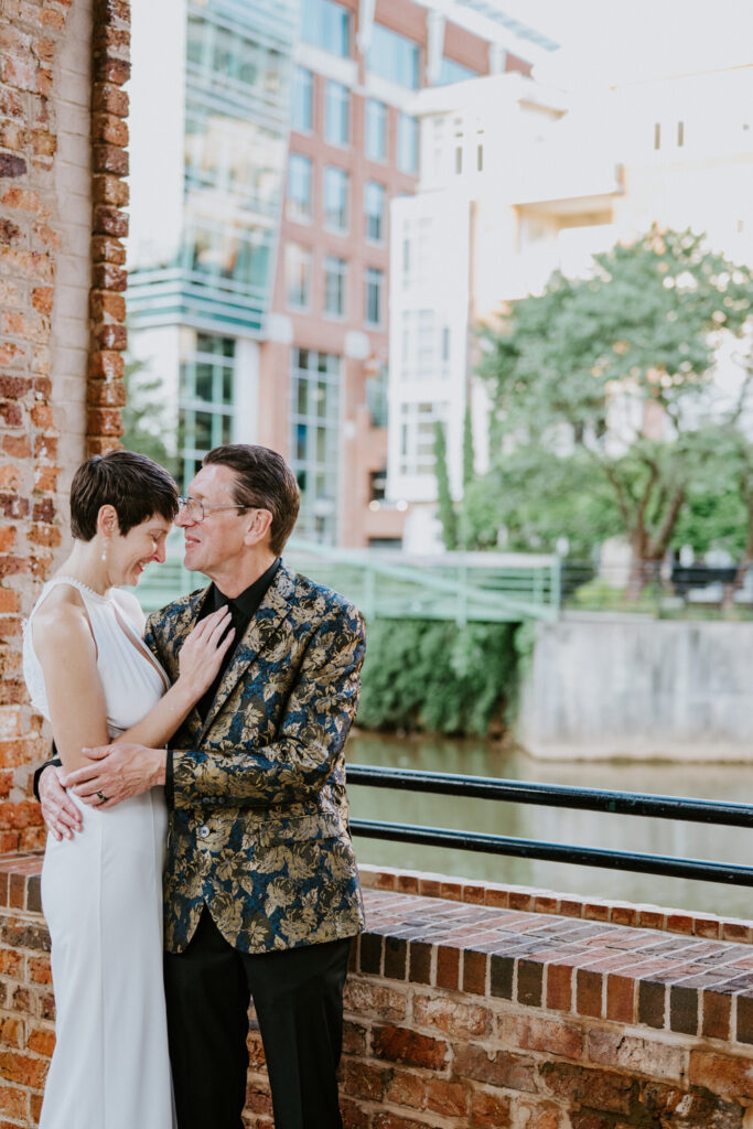 A couple stands lovingly embracing one another outdoors by a brick wall.