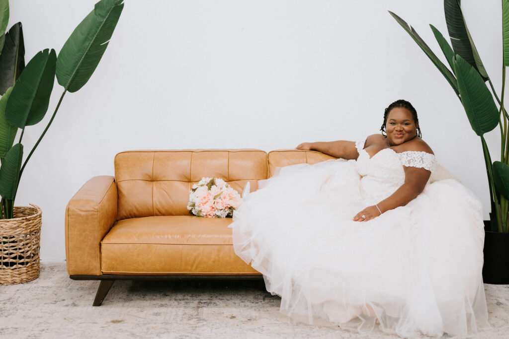 A beaming bride reclines on a caramel leather couch, her elegant lace wedding gown spread around her, with a bouquet of peach flowers in her hand, all framed by lush green tropical leaves in a chic, bright setting.