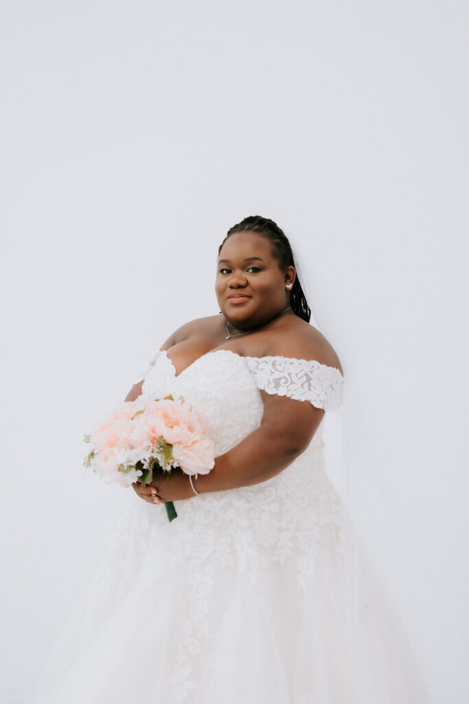 A radiant bride in a lace off-the-shoulder wedding gown smiles joyfully, clutching a bouquet of soft pink flowers, set against a white background that enhances the elegance of the moment.