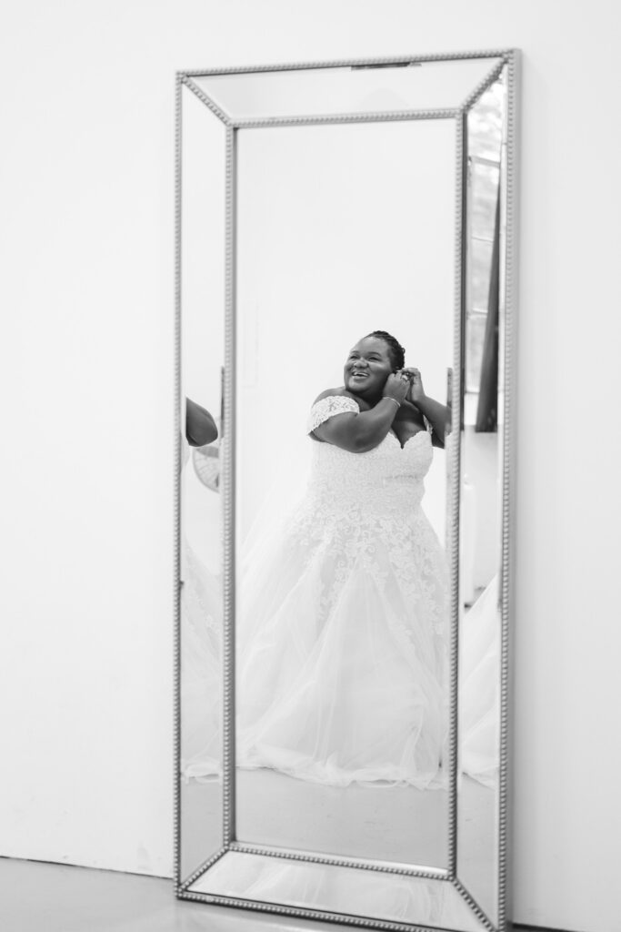 A beautiful monochrome shot of a bride in a mirror, her happiness is evident as she adjusts her earring, with the wedding dress detailing in sharp focus, reflecting a moment of anticipation.