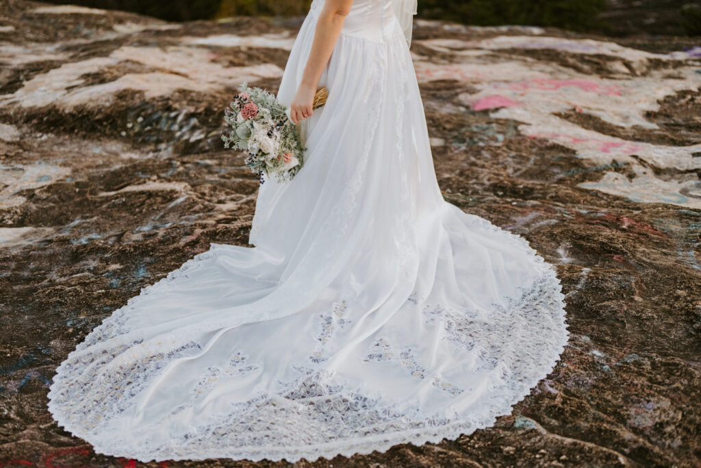A bride stands on a rocky terrain, her exquisite wedding gown train with detailed lace patterns trailing behind her, and a bouquet of rustic flowers in her hand, melding the elegance of the dress with the rugged beauty of nature.
