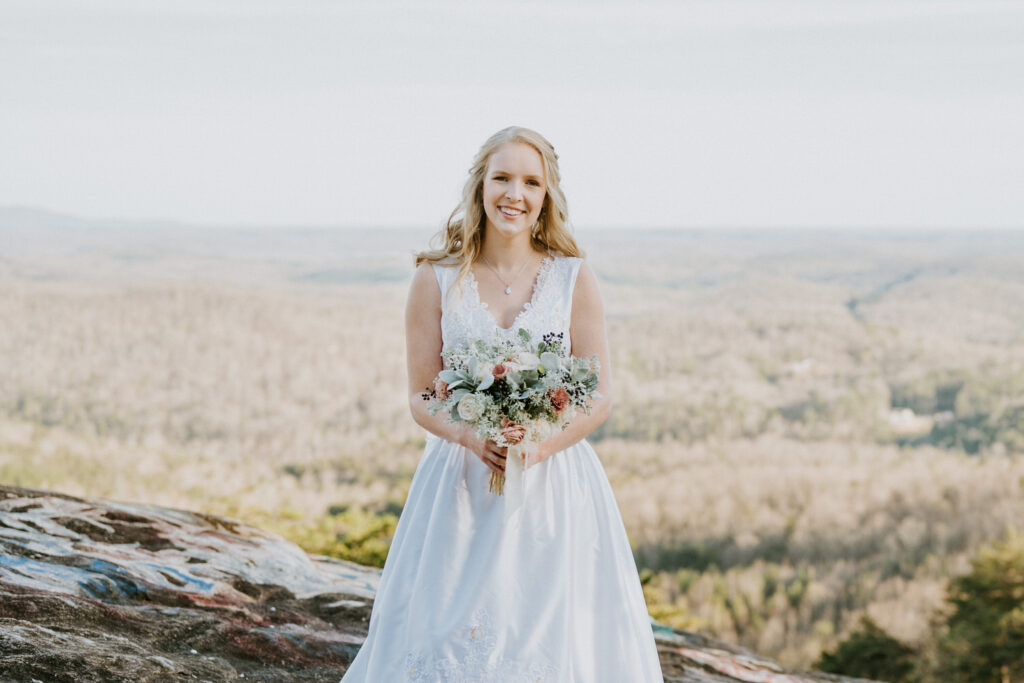 Smiling bride in a V-neck lace wedding gown holds a rustic bouquet, standing on a rocky outcrop with a vast forest landscape stretching into the horizon behind her.