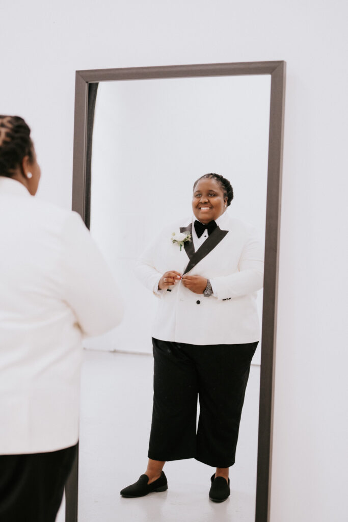 The bride, with a heartfelt smile, fixes her boutonniere while reflecting on her image in the mirror, surrounded by the simplicity of the room's decor.