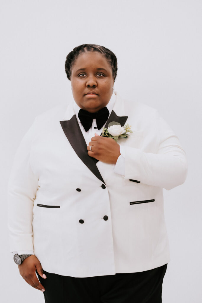 A portrait of a bride in a white tuxedo jacket with a boutonniere, her expression poised and confident, against a minimalistic backdrop.