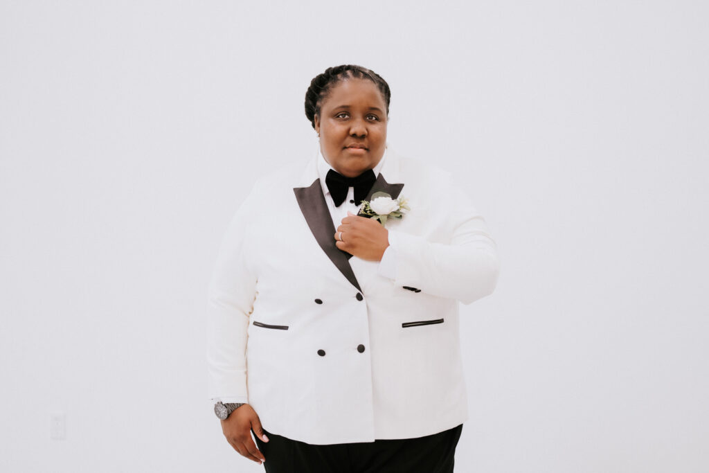 A poised individual in a white double-breasted tuxedo jacket with black lapels and buttons, holds a boutonniere, exuding elegance against a white backdrop.