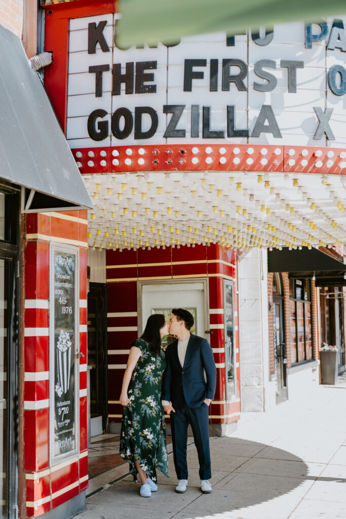 A man and a woman, both Korean, hold hands. They are standing under the marquee for a movie theater. They are leaning in to kiss. The man is wearing a navy suit and the woman is wearing a long green floral dress.
