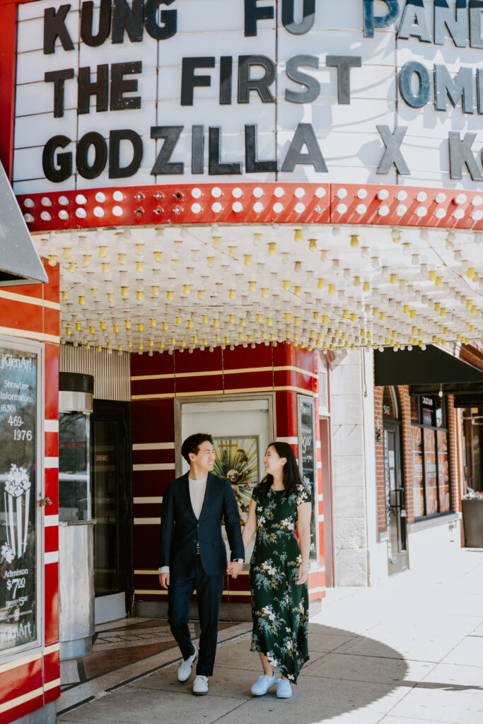 A man and a woman, both Korean, hold hands. They are standing under the marquee for a movie theater. They are smiling at each other. The man is wearing a navy suit and the woman is wearing a long green floral dress.