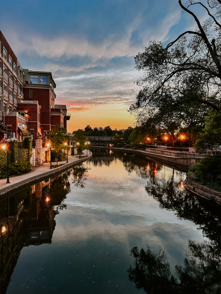 A view of the river winding through downtown Naperville at sunset. The streetlights have just come on and the sky is oranges and blues.