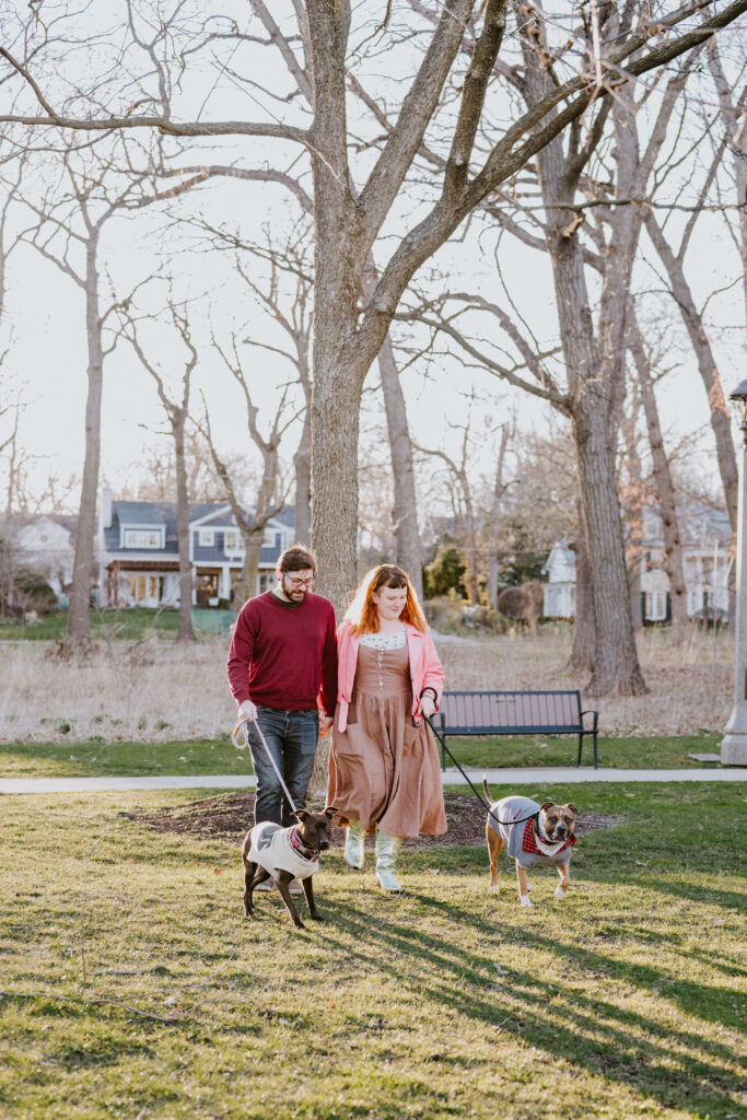 A man in a red sweater and a woman in a brown dress walk through a park. The man has a small black dog in a white sweater on a leash, and the woman has a larger brown dog in a gray sweater on a leash.