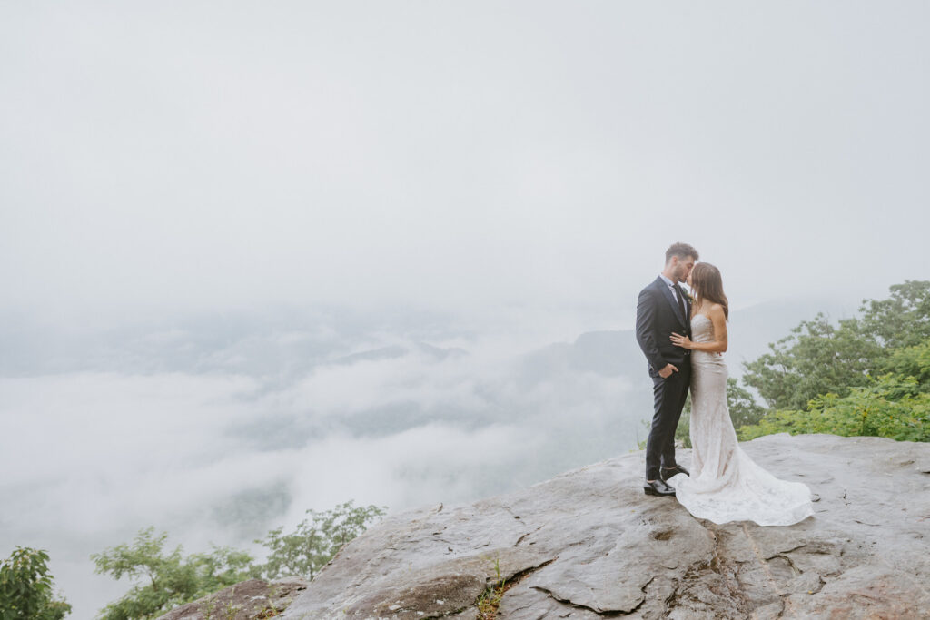 A couple kisses on a rock with mountains behind them.